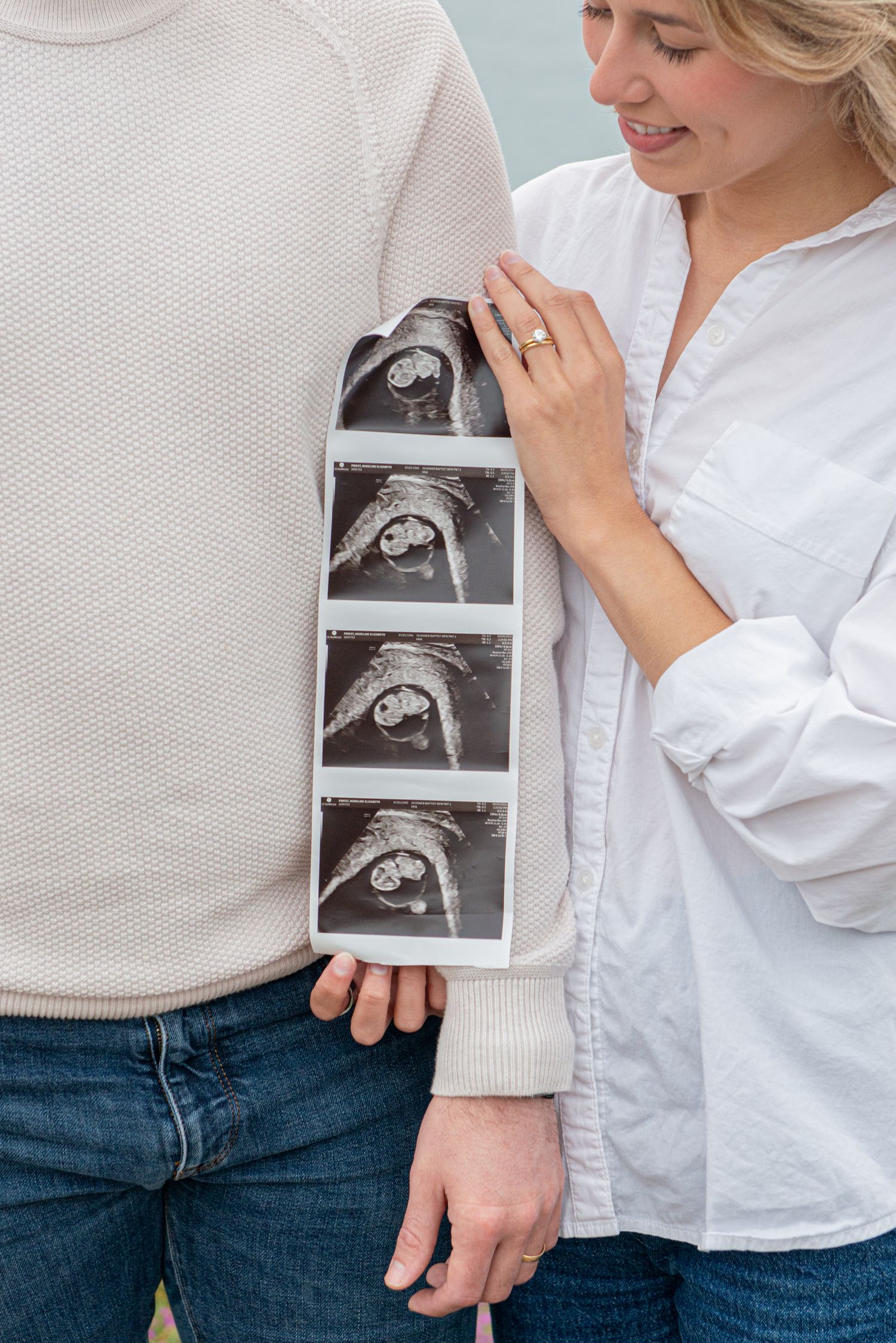 Smiling pregnant woman in white shirt holding ultrasound sonogram strip beside her partner.
