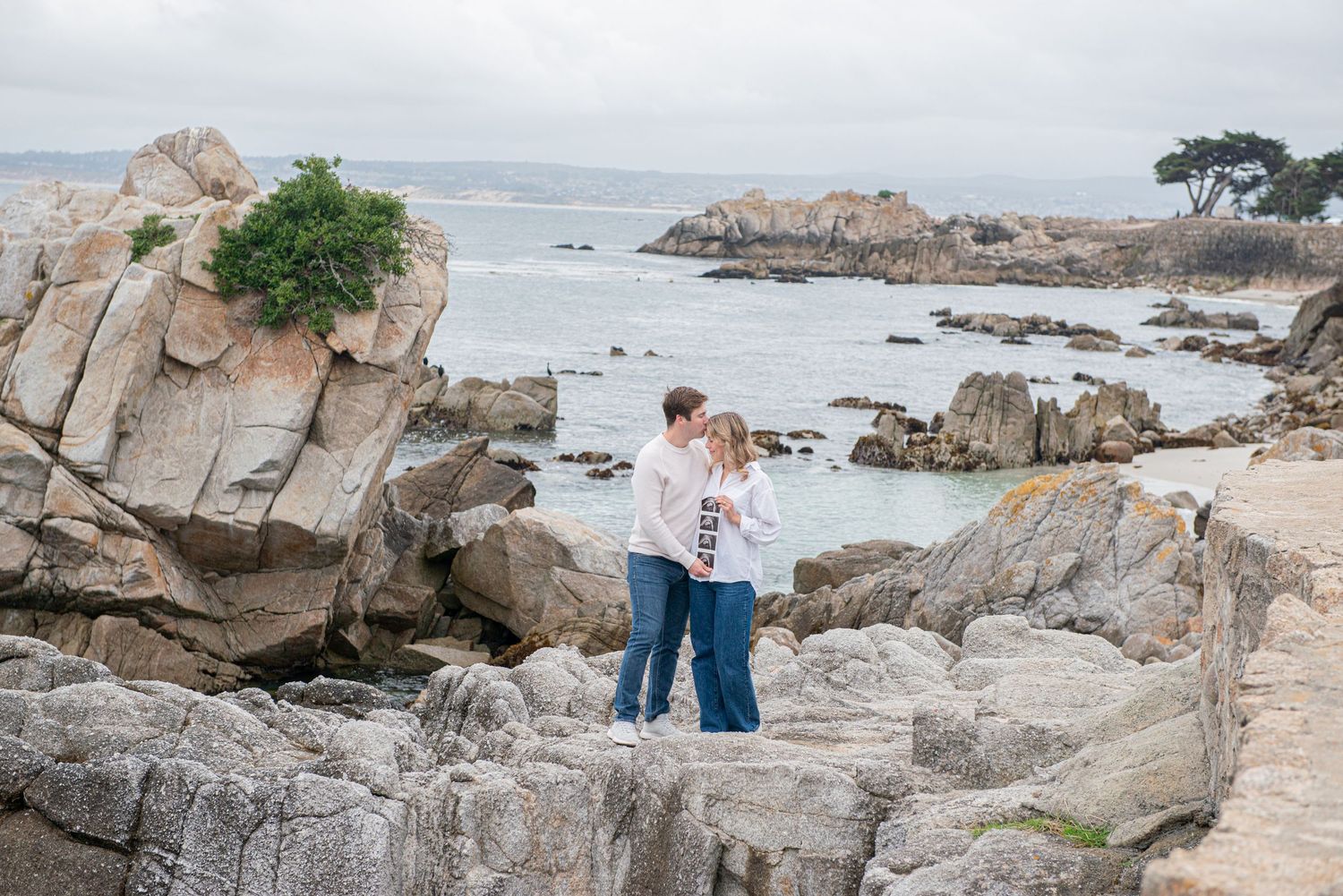 Couple embracing on rocky coastal shoreline with ocean waves and boulders in the background on a cloudy day.