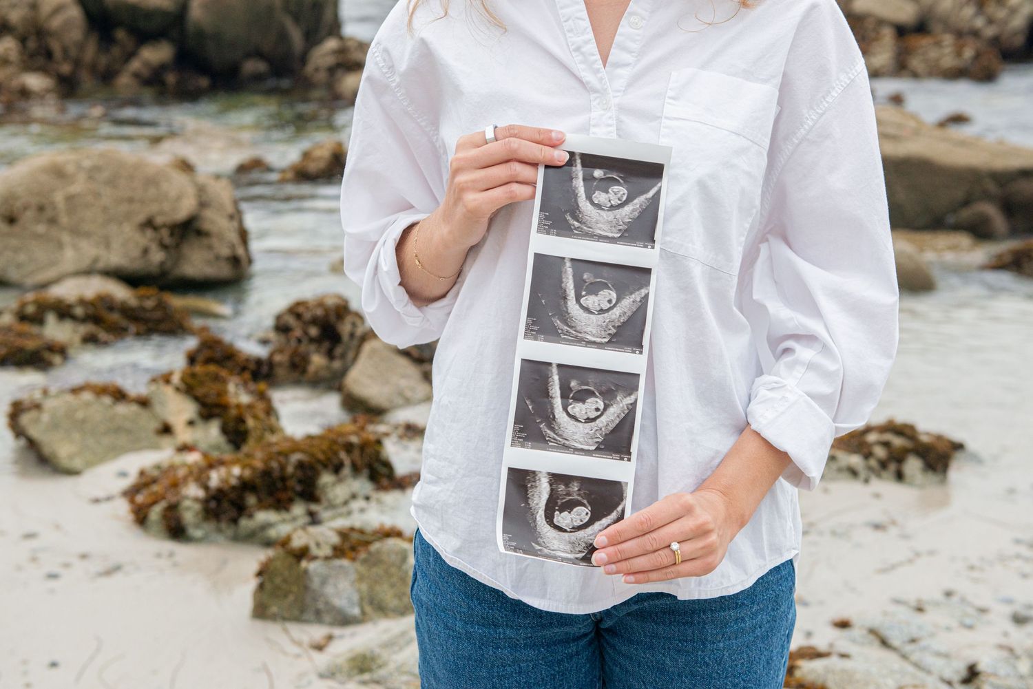 Pregnant woman in white shirt holding ultrasound strip photos at rocky beach, showing baby sonogram images.