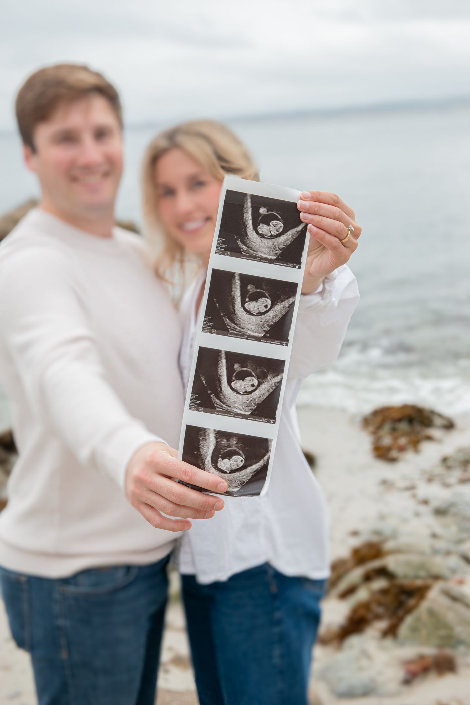 Happy couple holding up baby ultrasound sonogram strip at a rocky ocean beach.