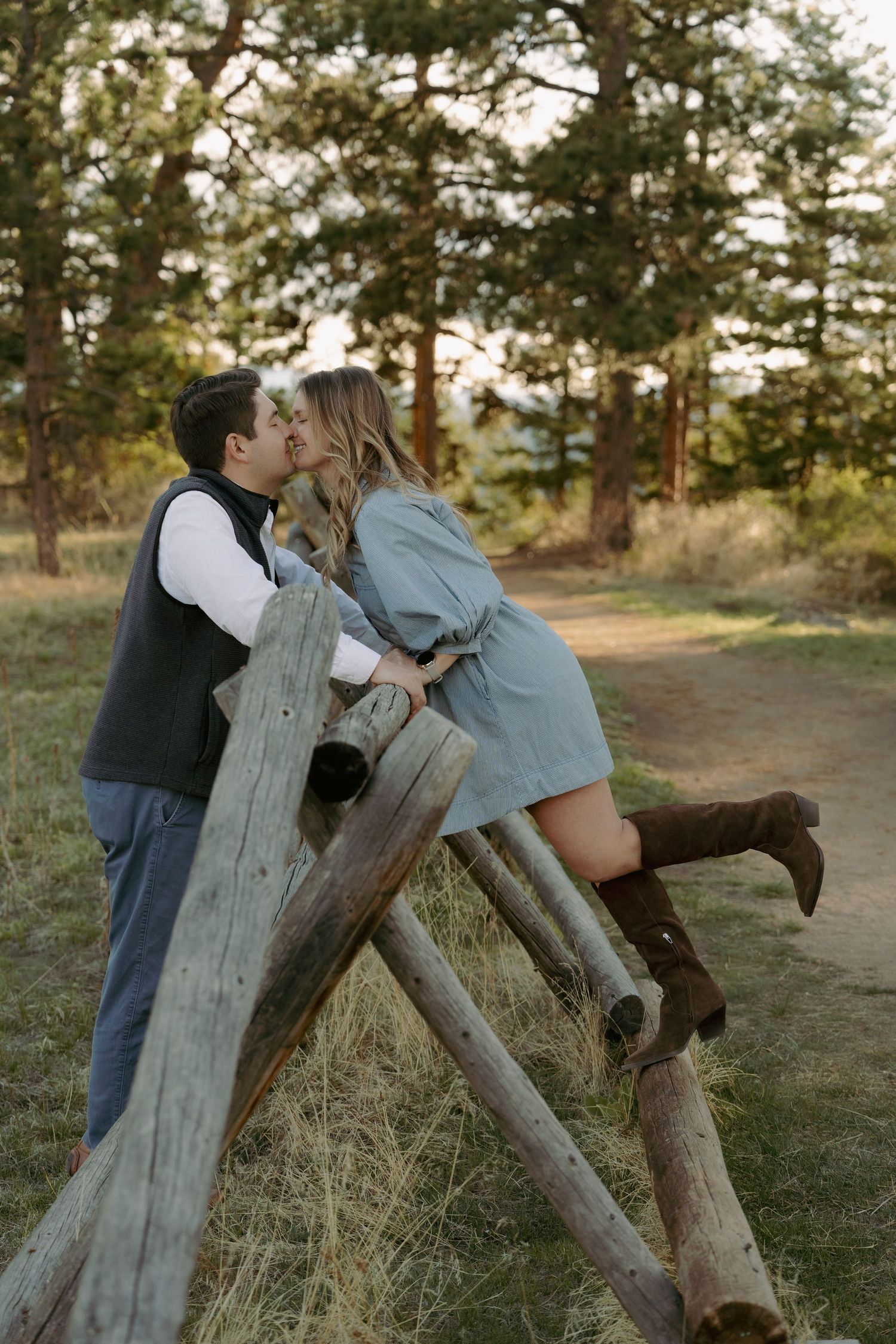 Couple sharing a romantic kiss leaning on a rustic wooden fence in a scenic forest setting, woman wearing brown boots.