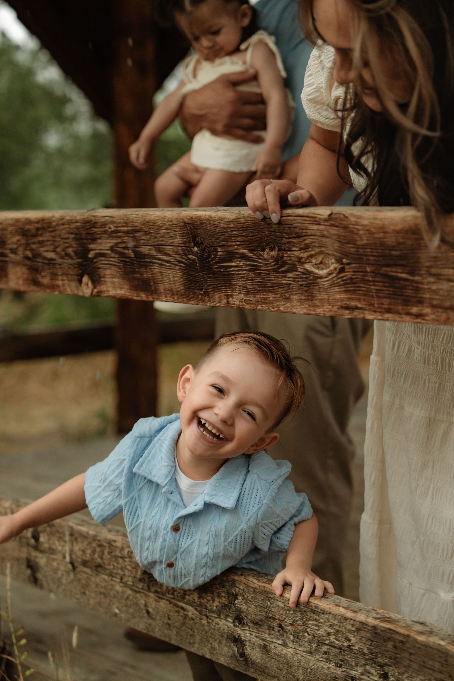 A laughing toddler in a blue shirt peeks through wooden fence rails while a woman holds a baby above.