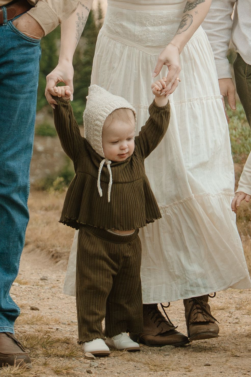 A baby in olive ribbed outfit and bonnet walks outdoors, held by parents in jeans and white dress.
