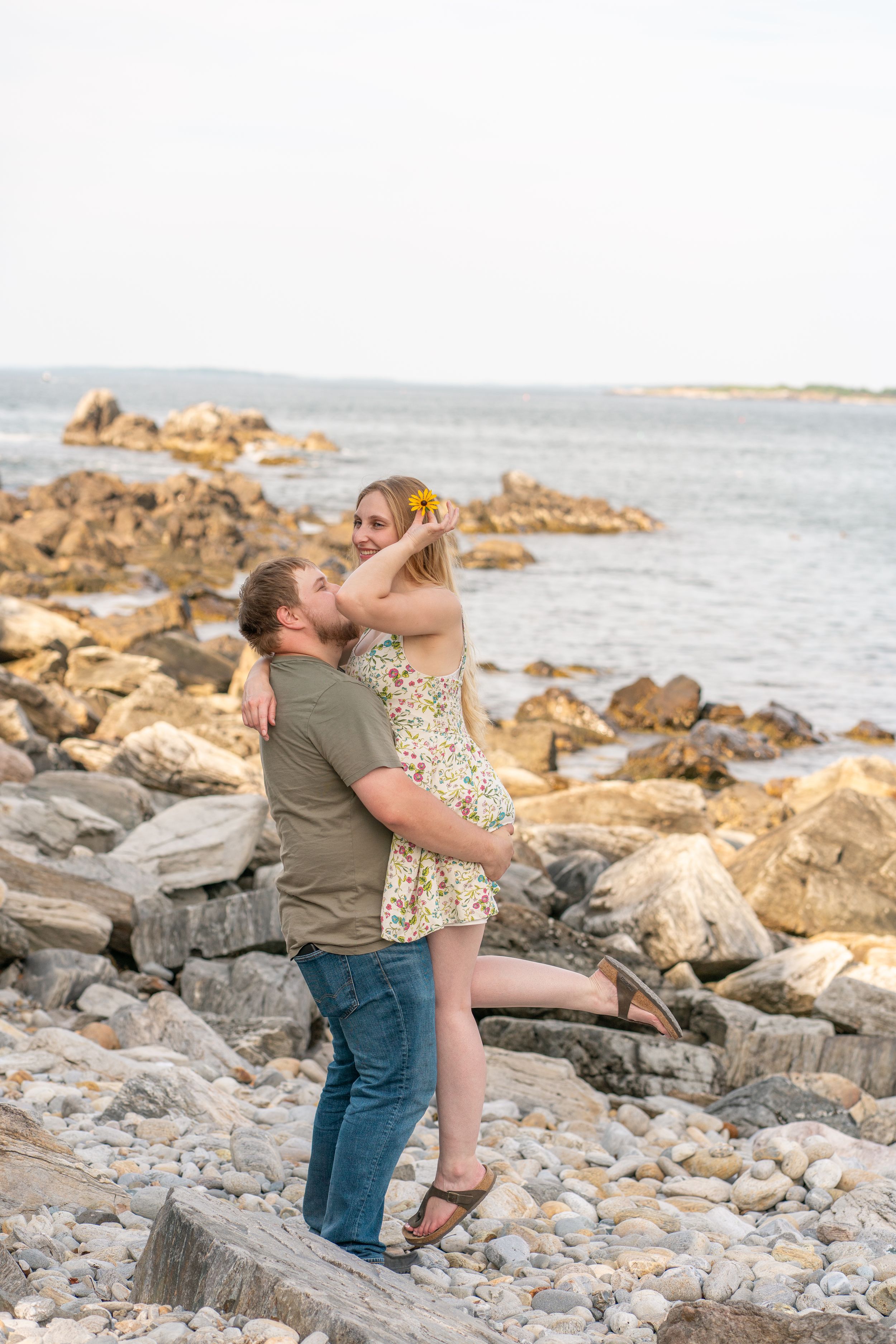 Romantic couple embracing on rocky beach shoreline, man lifting woman in floral dress at sunset.