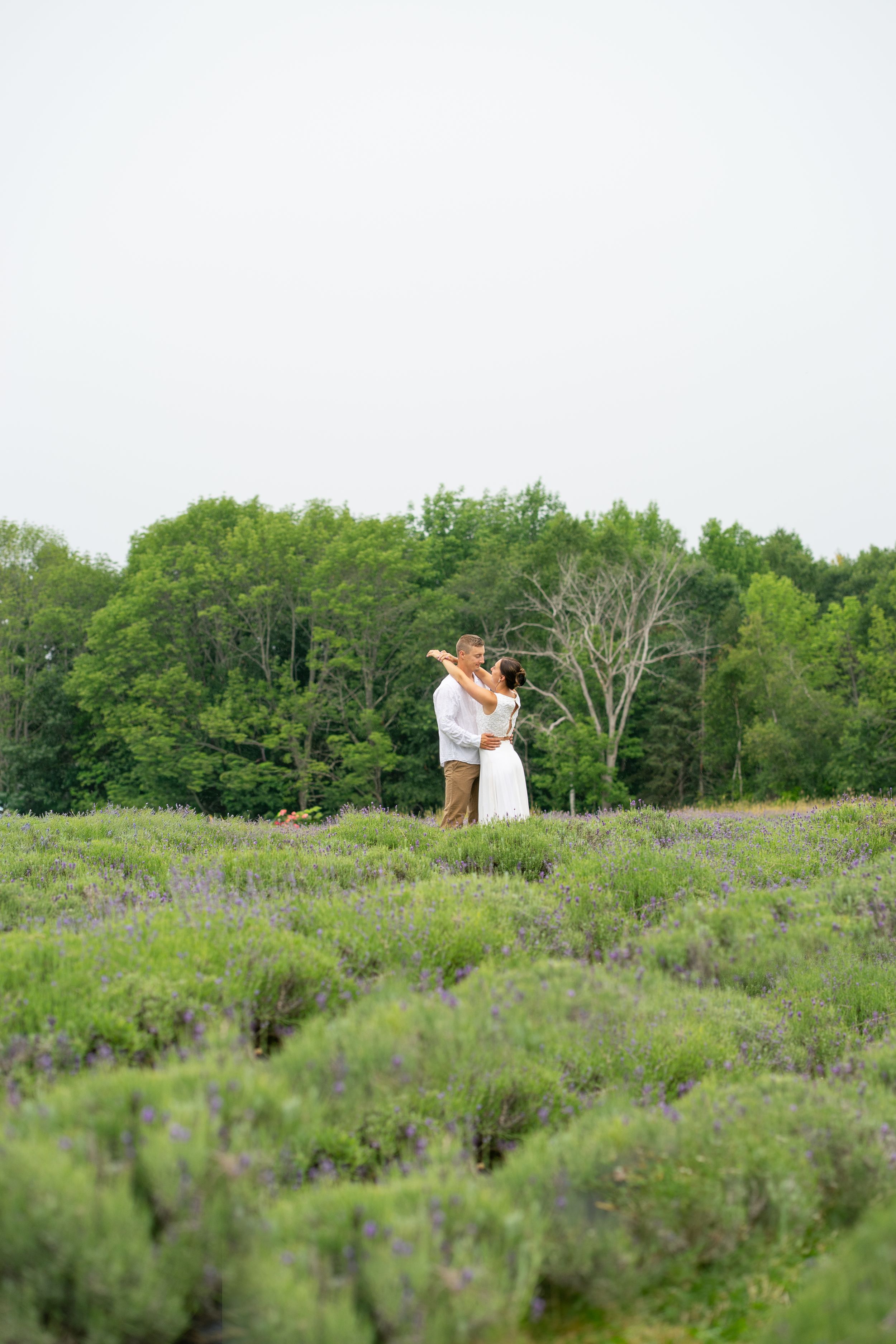 A couple embraces and dances together in a vast lavender field surrounded by lush green trees.