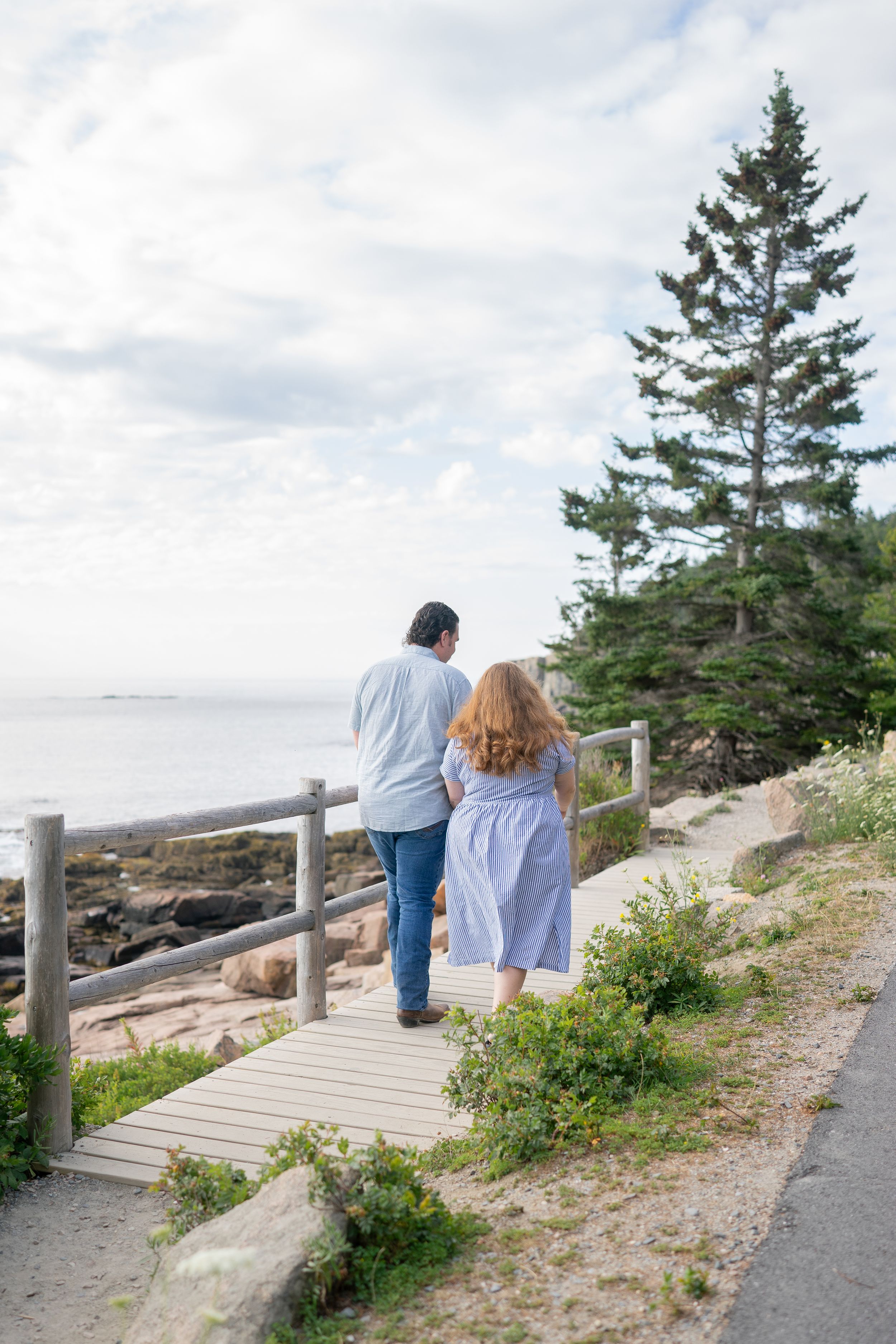 Couple walking hand in hand on a wooden boardwalk overlooking a rocky New England coastline.