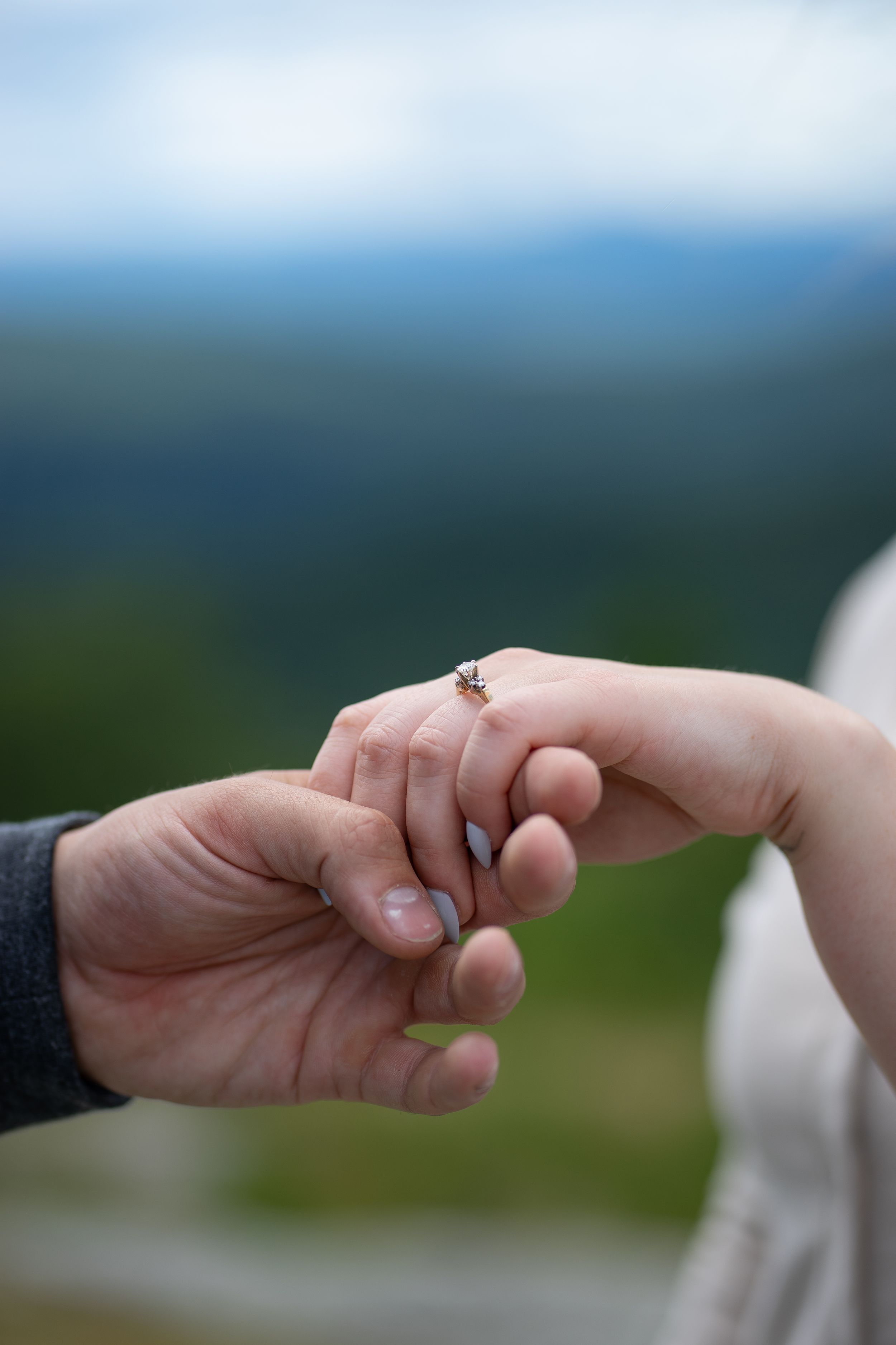 Close-up of couple holding hands with engagement ring on mountain backdrop.