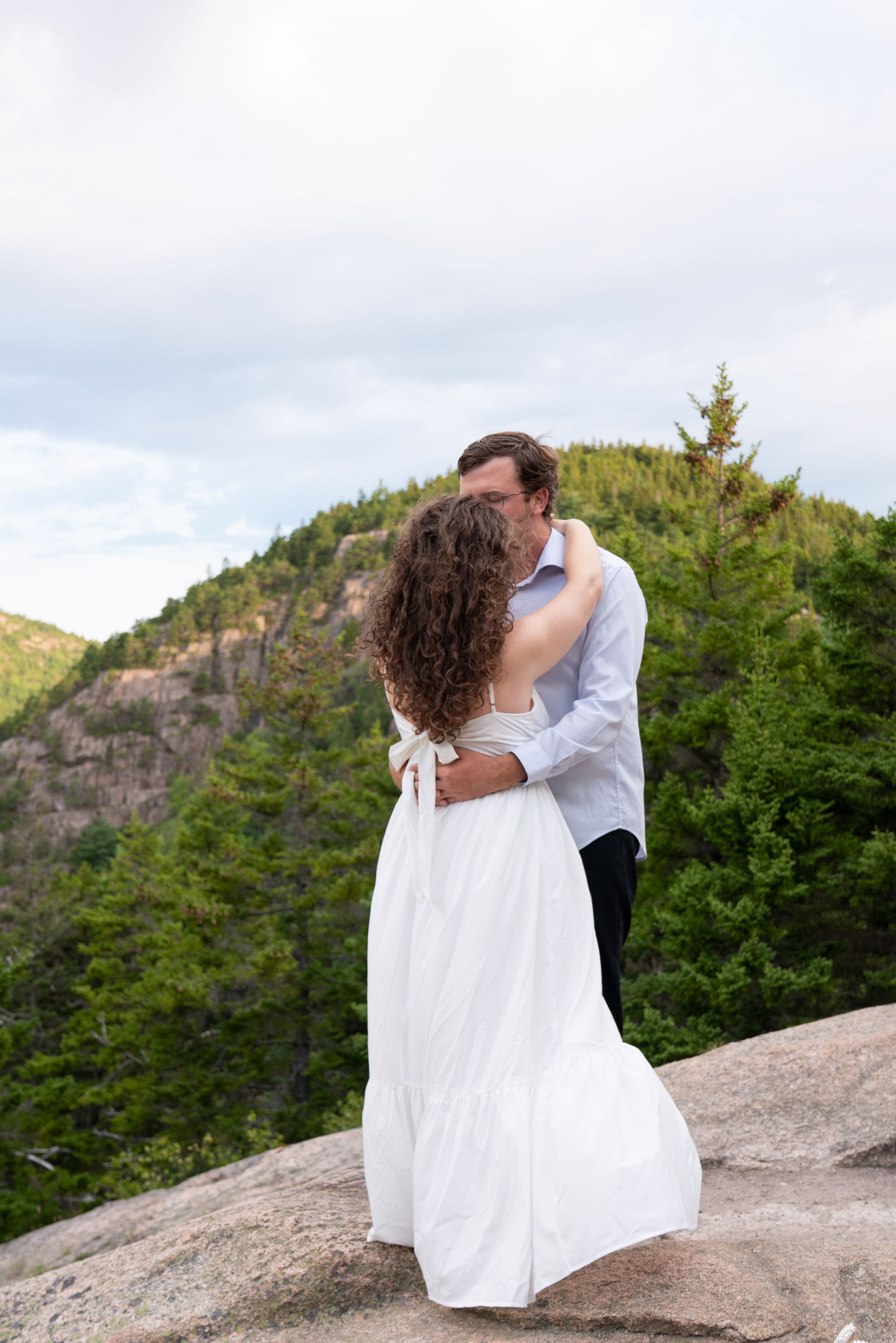 Couple sharing a romantic kiss on a rocky mountaintop, woman in white dress, surrounded by lush green forest and mountains.