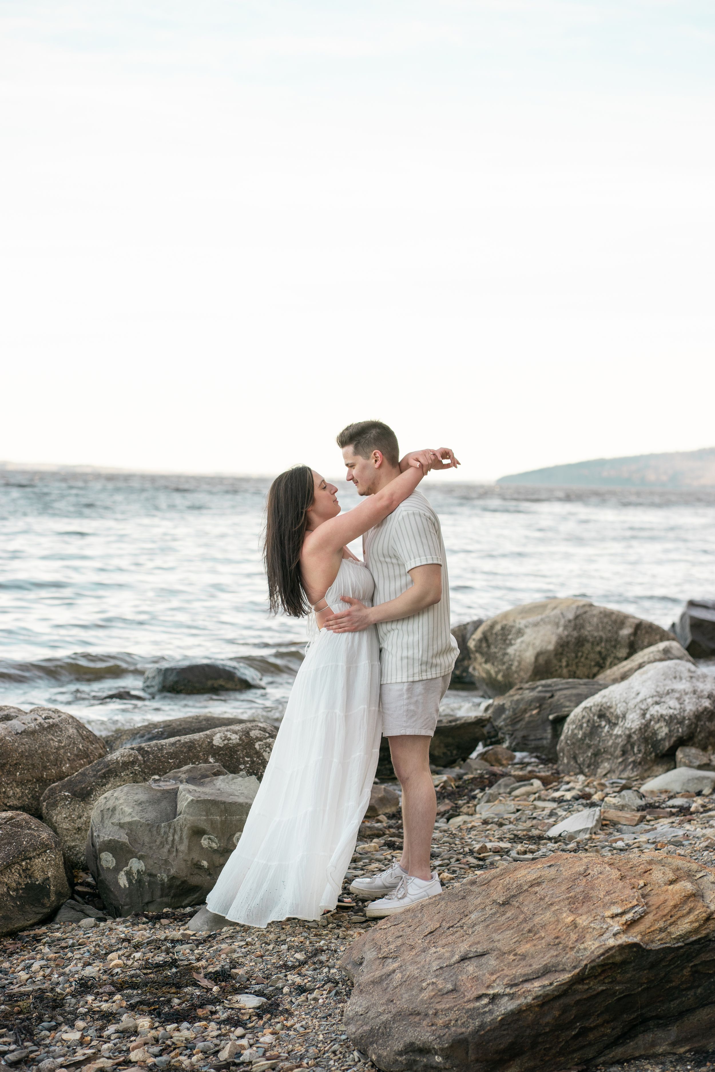 Romantic couple embracing on a rocky beach at sunset, woman in flowing white dress.