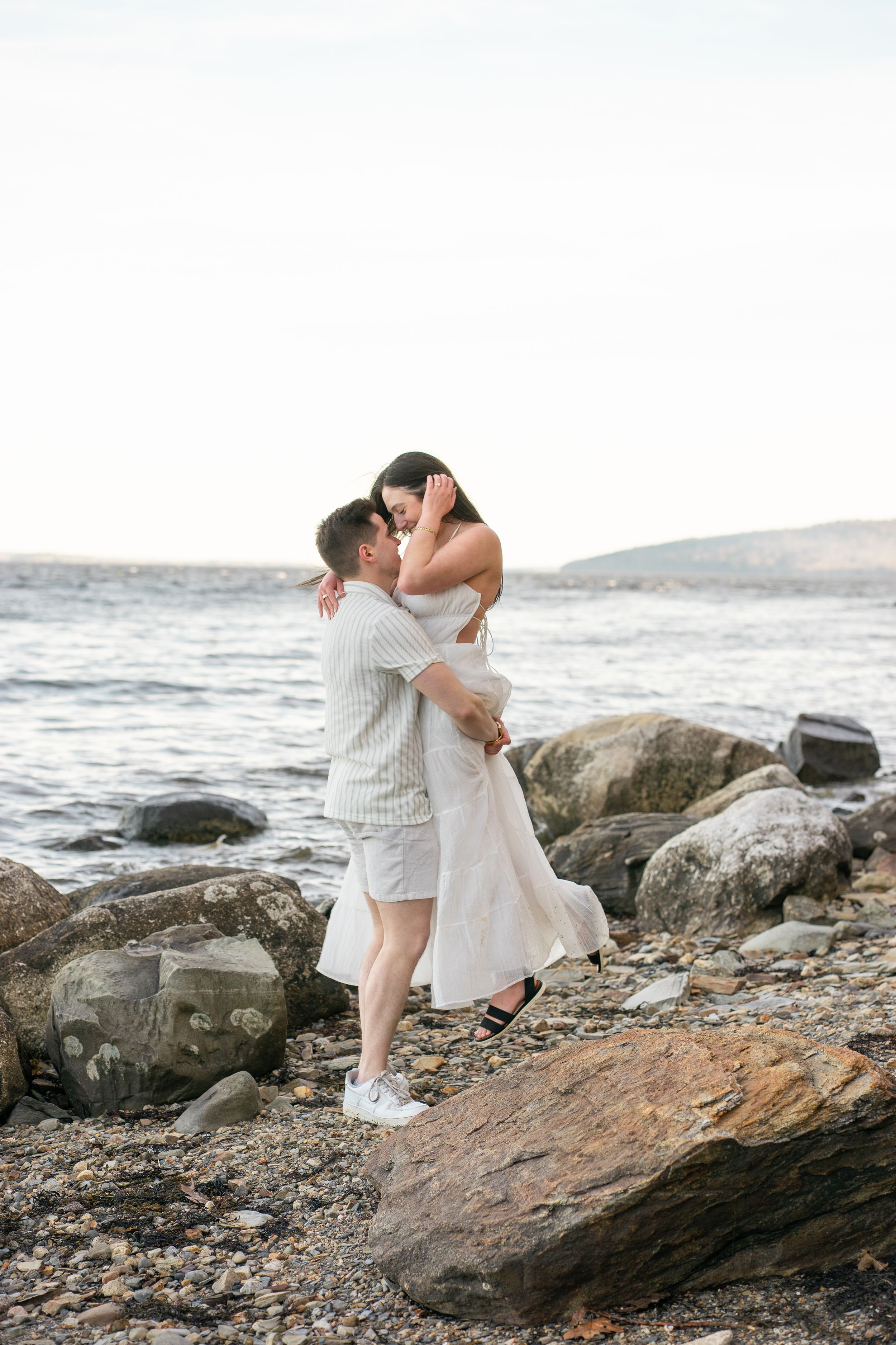 Man lifting woman in white dress on a rocky shoreline, both laughing joyfully.