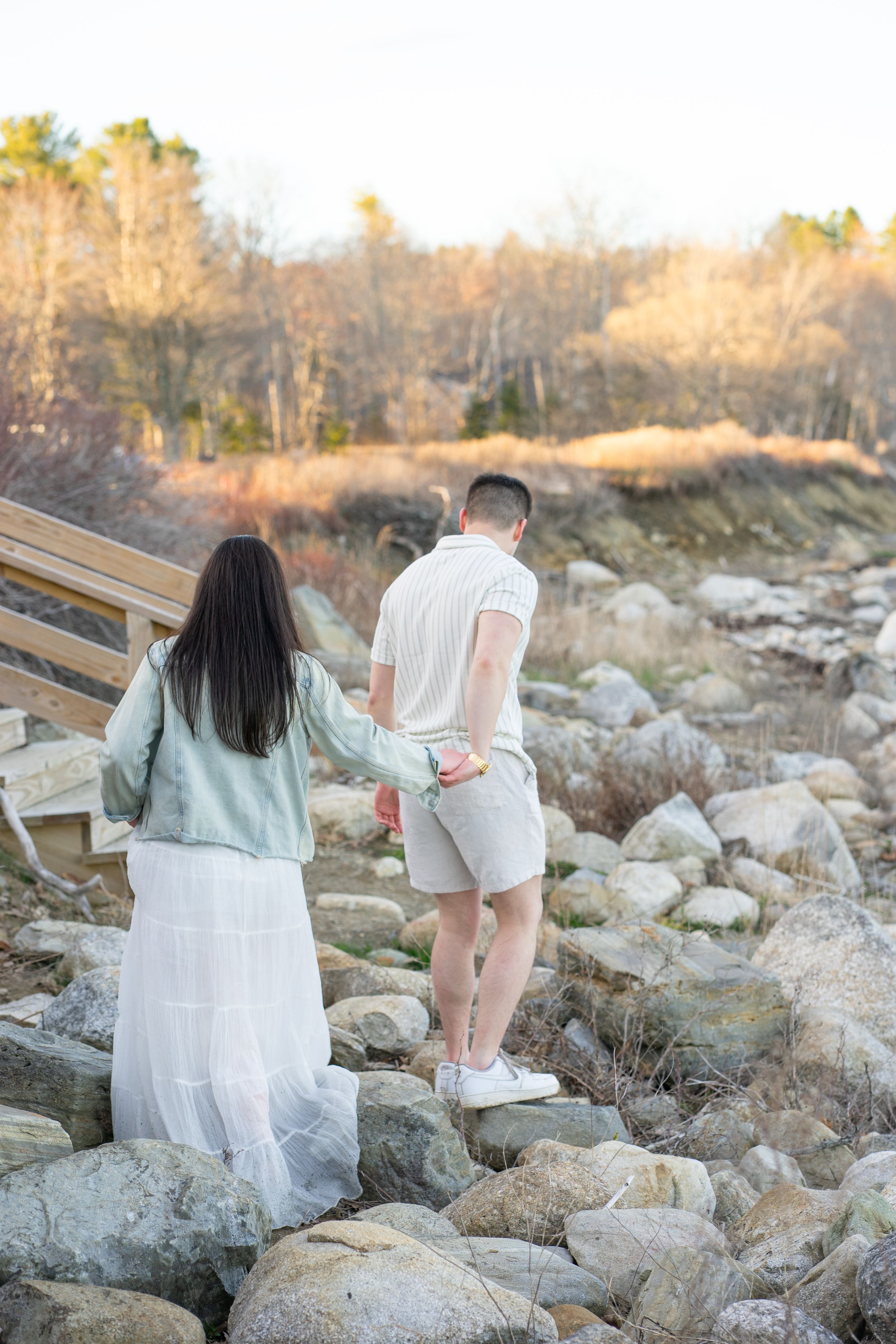 Couple holding hands walking over rocks near a wooden staircase at golden hour.