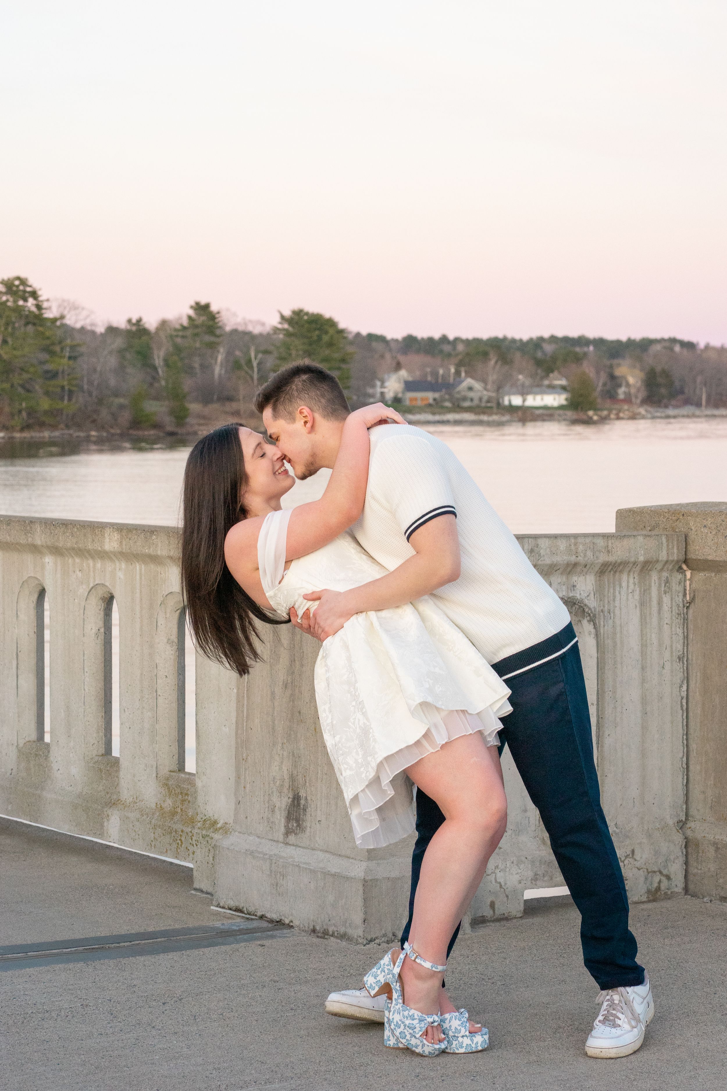 Couple shares romantic dip kiss on stone bridge at sunset by water.