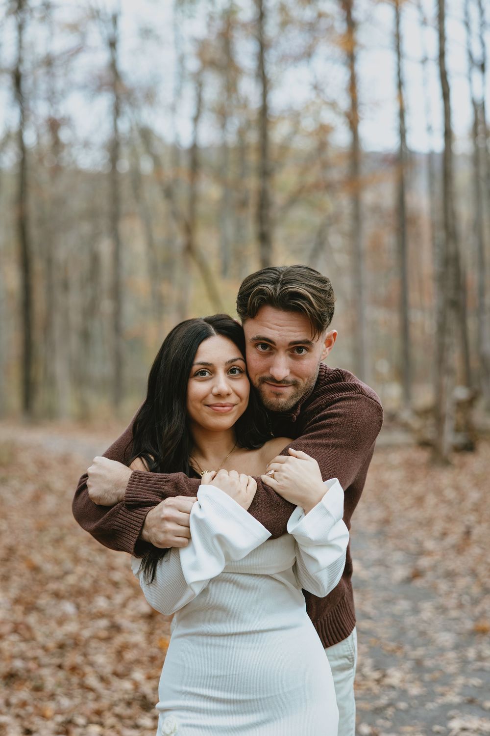 Couple embracing in autumn forest, woman in white dress, man in brown sweater, surrounded by fall foliage.