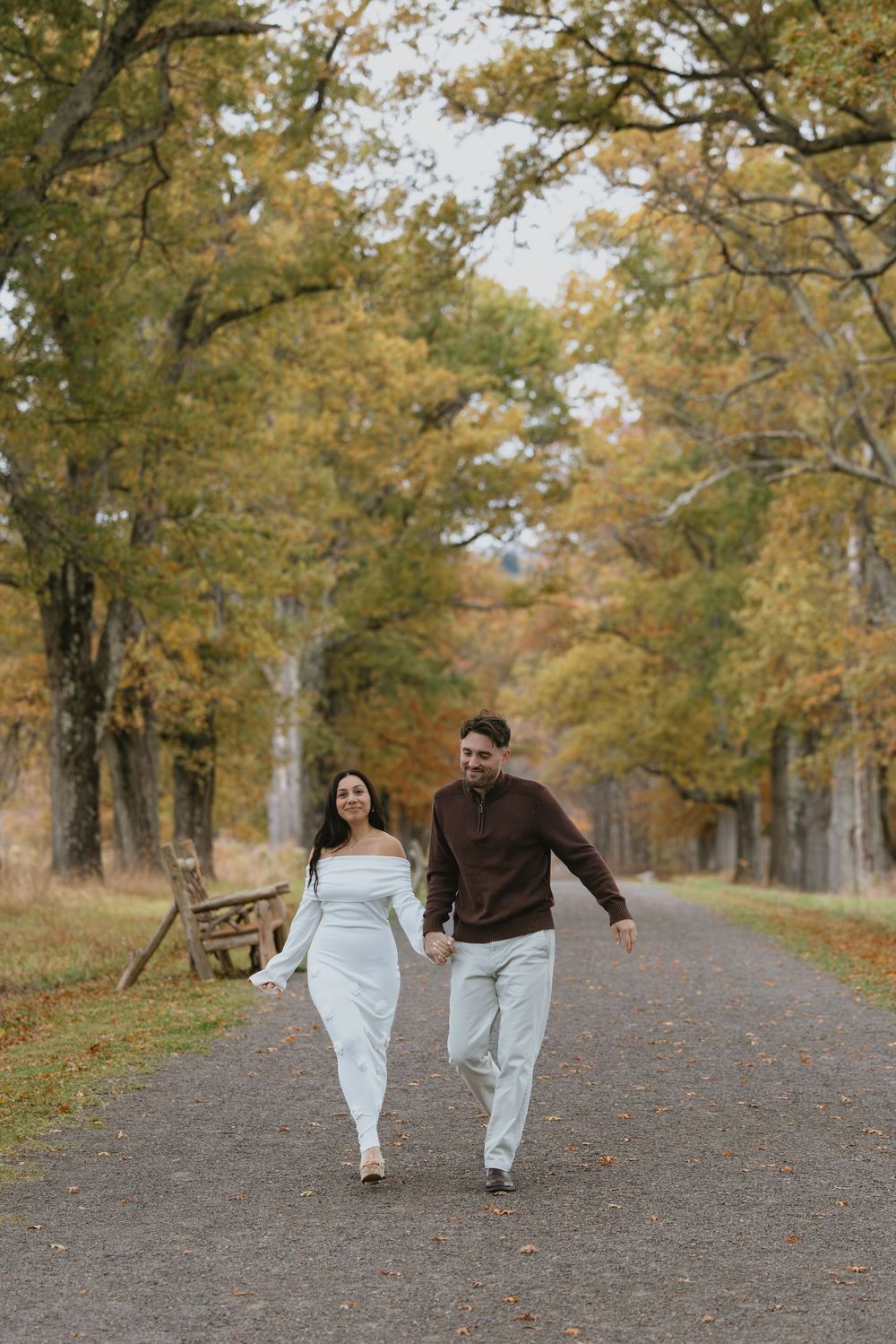 Couple holding hands walking down a tree-lined path surrounded by colorful autumn foliage.