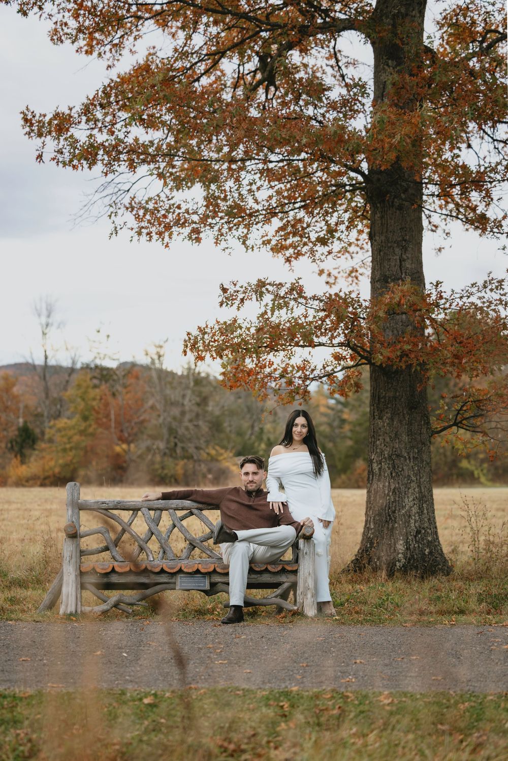 Couple posing by a wooden bench under a large autumn tree with colorful foliage in a scenic outdoor park setting.