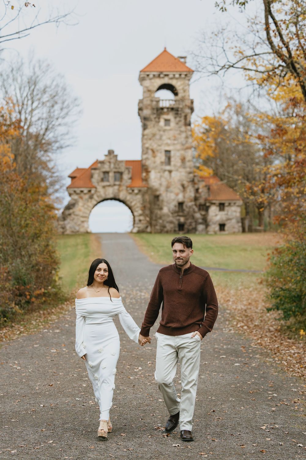 Couple holding hands walking on a path toward a stone castle tower surrounded by autumn foliage.