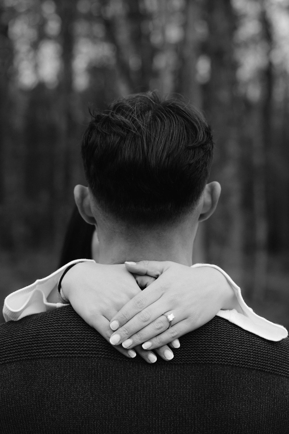 Black and white photo of a couple embracing outdoors, woman's hands with ring wrapped around man's neck.