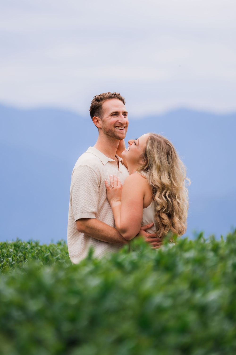 Smiling couple embracing in a lush green field with mountain backdrop during an outdoor engagement photo session.