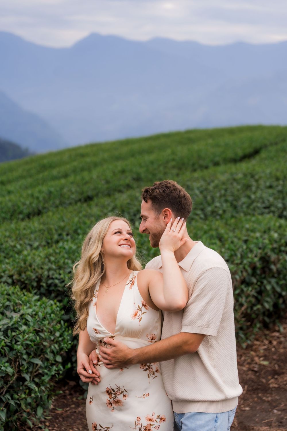 Couple embracing and smiling at each other in a lush green tea plantation with misty mountains behind.