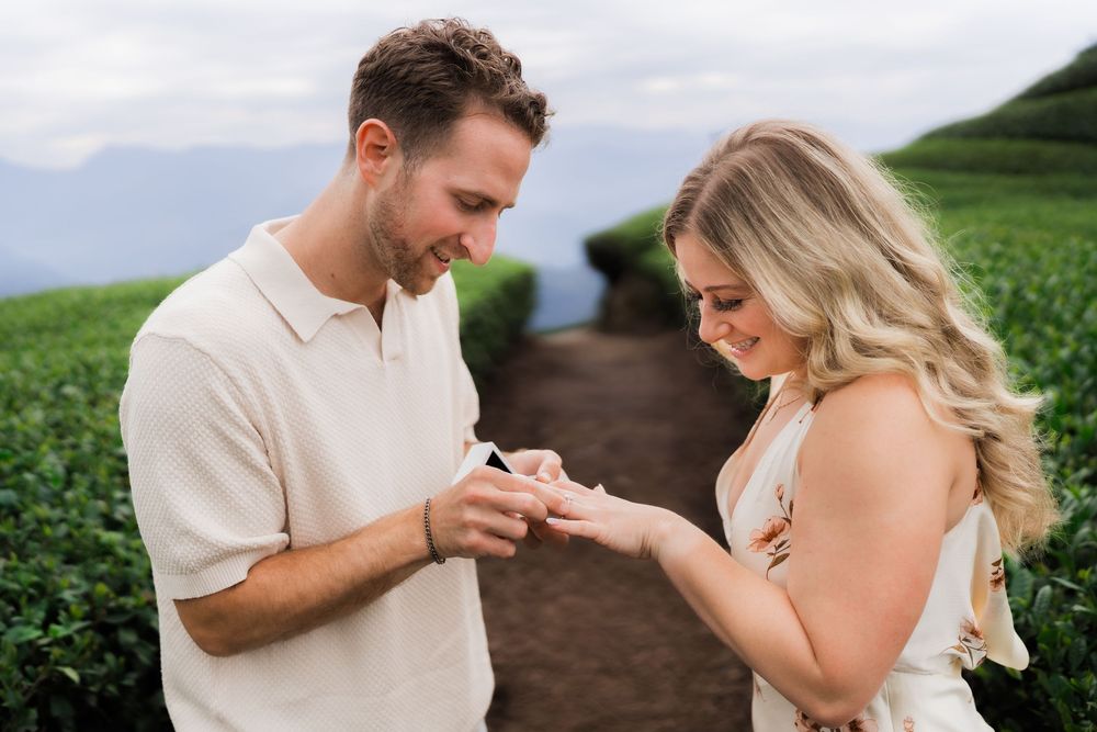 Man placing engagement ring on womans finger at a scenic green tea farm pathway.