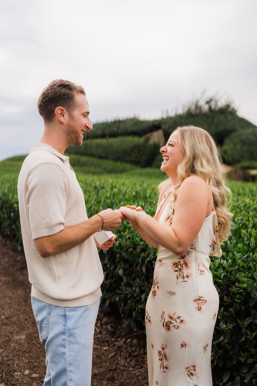 Smiling couple holding hands in a lush green tea plantation, woman in floral dress laughing joyfully.