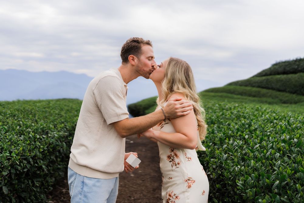 Couple sharing a romantic kiss in a lush green tea plantation field with mountains in the background.