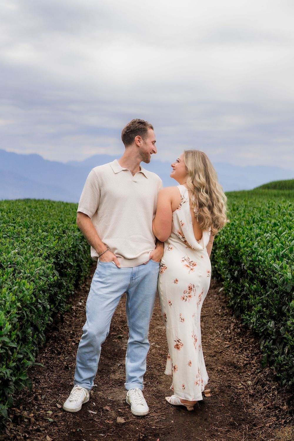 Couple smiling at each other in a lush green field with mountain views, man in blue jeans and woman in floral dress.