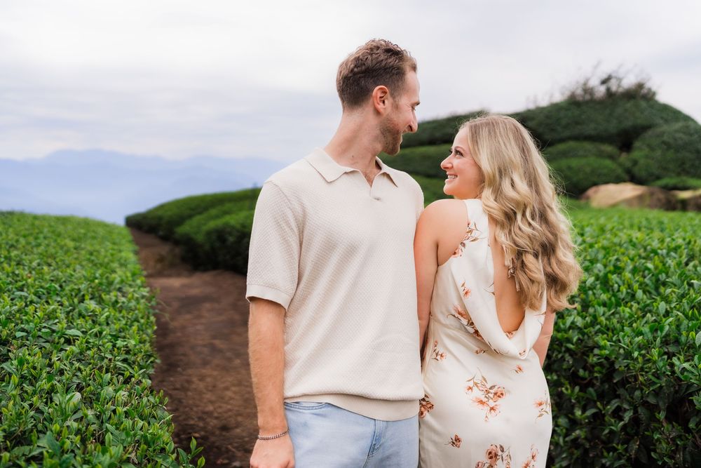 Couple smiling at each other in lush green tea plantation fields with misty mountains in background.