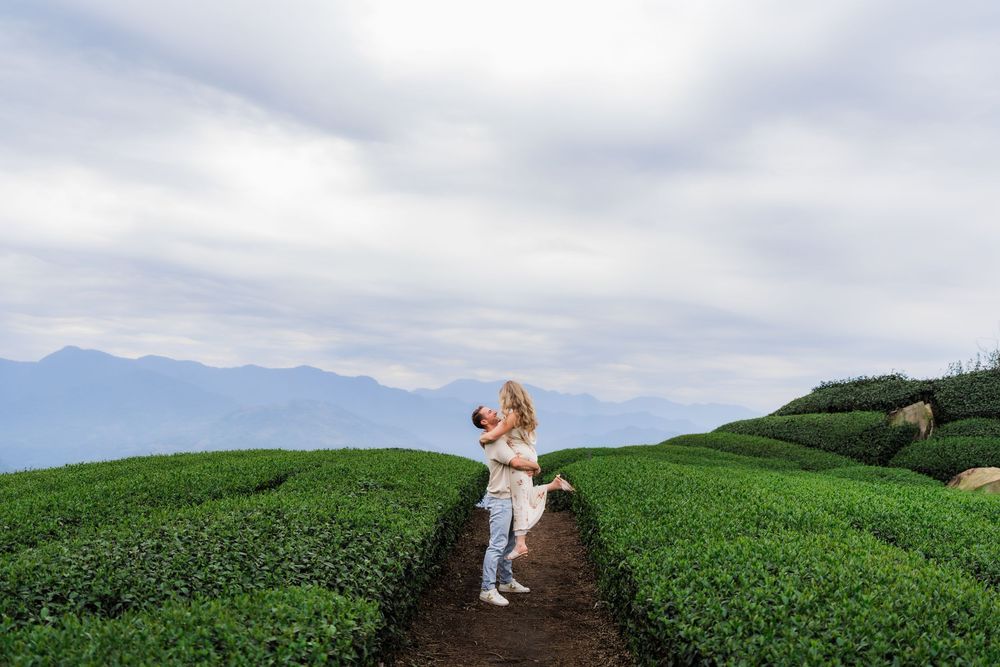 Man lifting woman joyfully on a path through green tea fields with mountains in the background.