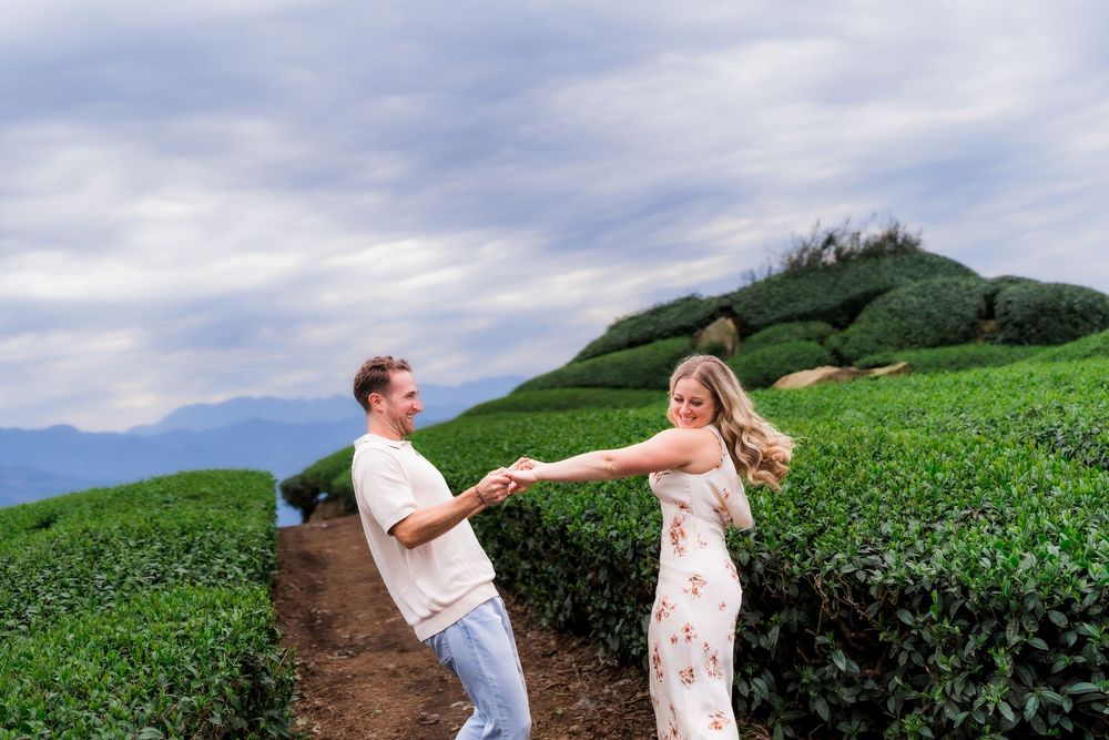 Joyful couple holding hands and spinning on a scenic tea plantation path with rolling green hills.