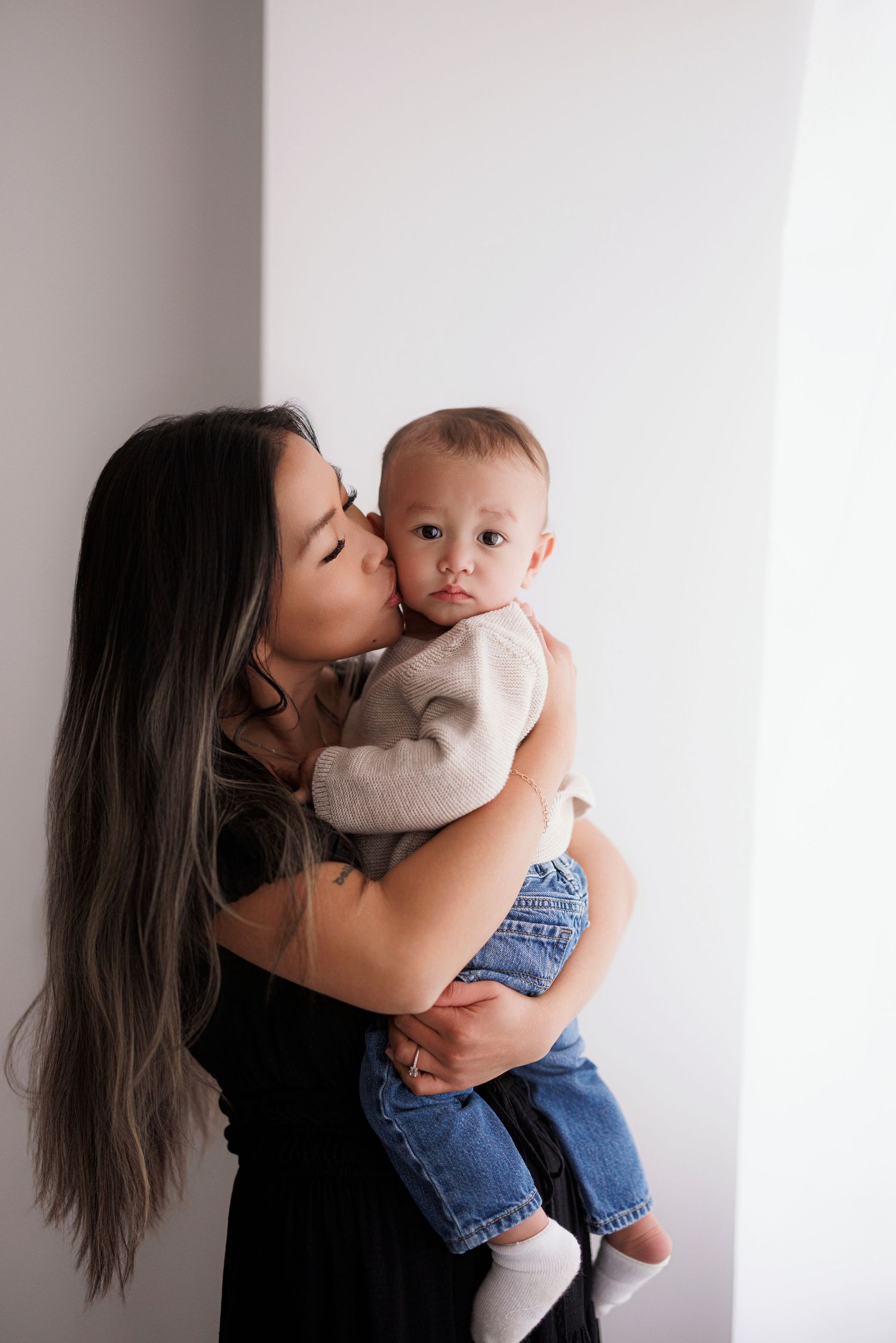A mother with long dark hair kisses her baby dressed in denim overalls against a white background.