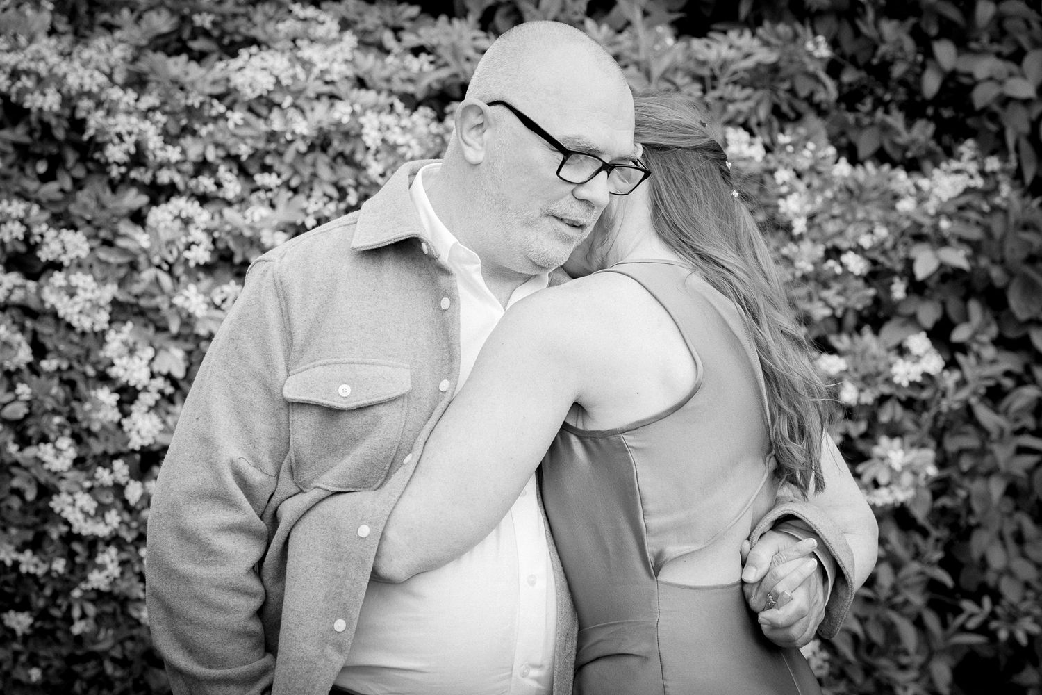 Black and white photo of a bald man with glasses embracing a woman outdoors near blooming flowers.