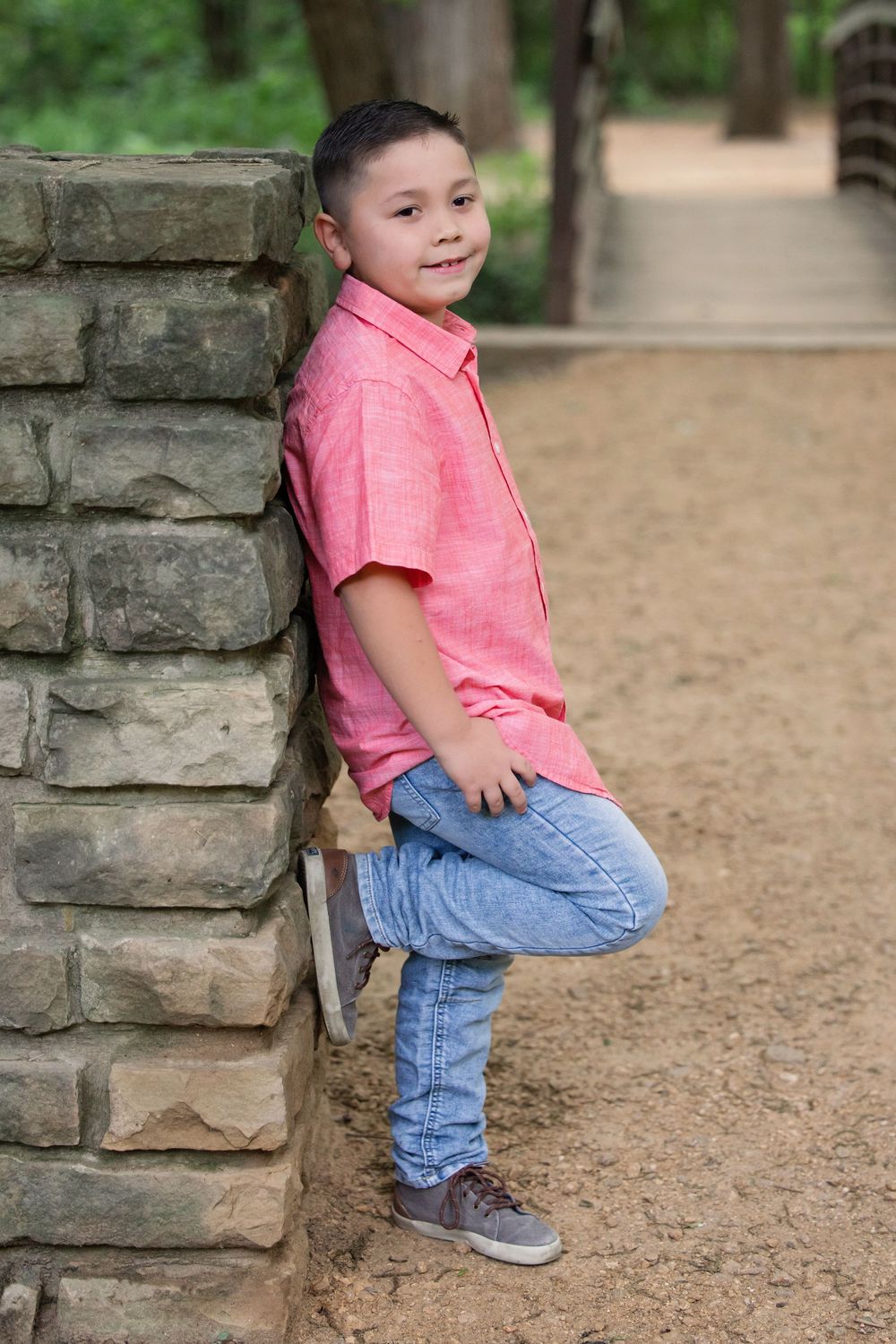 Young boy in pink shirt and jeans leans against stone pillar in outdoor park setting in Colleyville