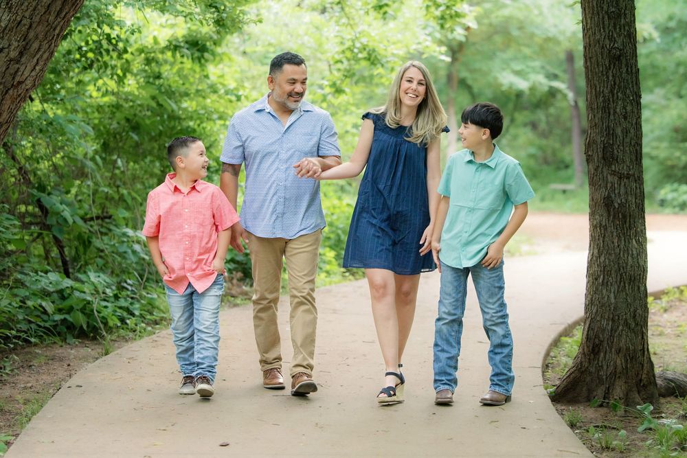 Family of four walking on a shaded park path, smiling and holding hands on a sunny day in Colleyville