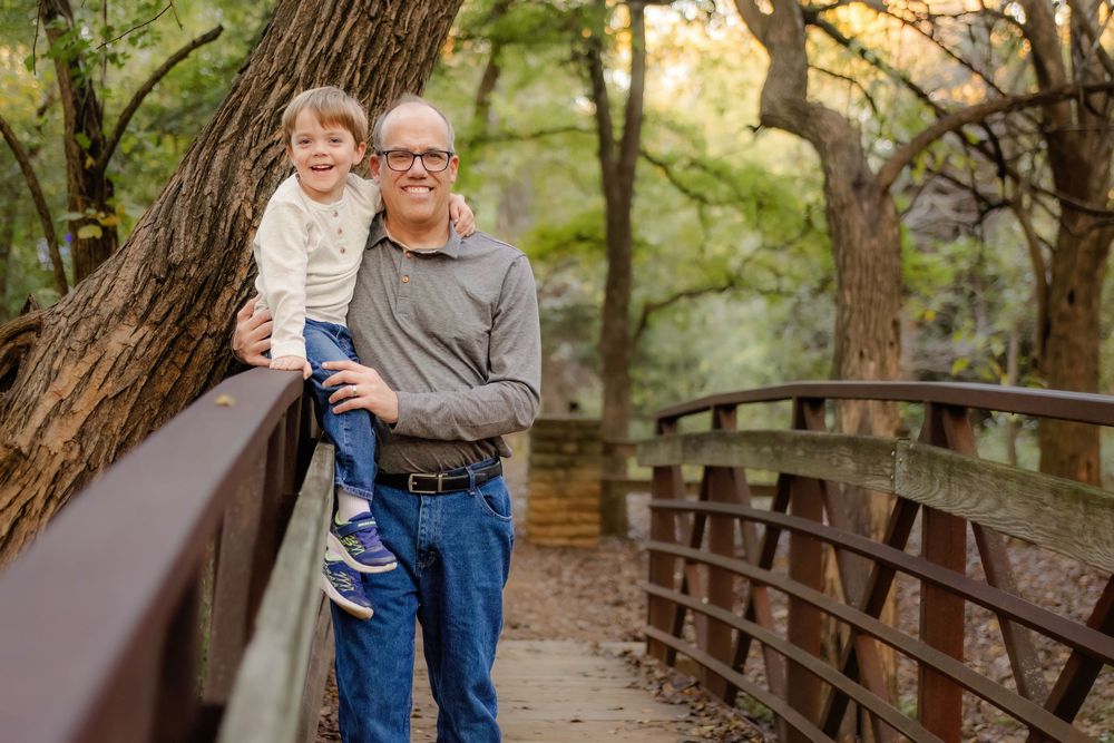 Man holding young child on a wooden bridge surrounded by lush green trees in a park setting in Colleyville.