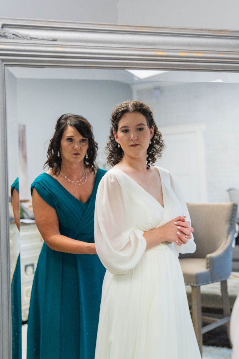 A bride in a white gown stands before a mirror as a bridesmaid in teal adjusts her dress.