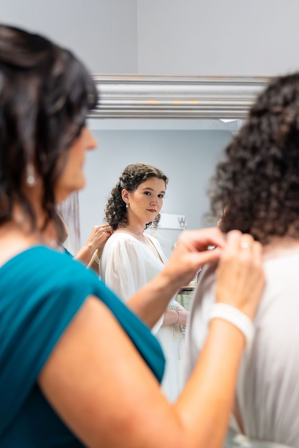 Bridesmaid helping bride with dress zipper while bride views reflection in mirror before wedding ceremony.