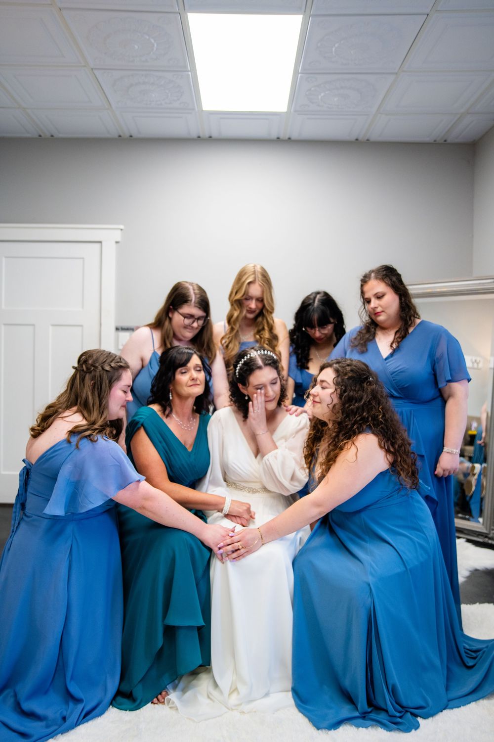 Bridesmaids in blue dresses gather around emotional bride in white gown for pre-wedding prayer circle.