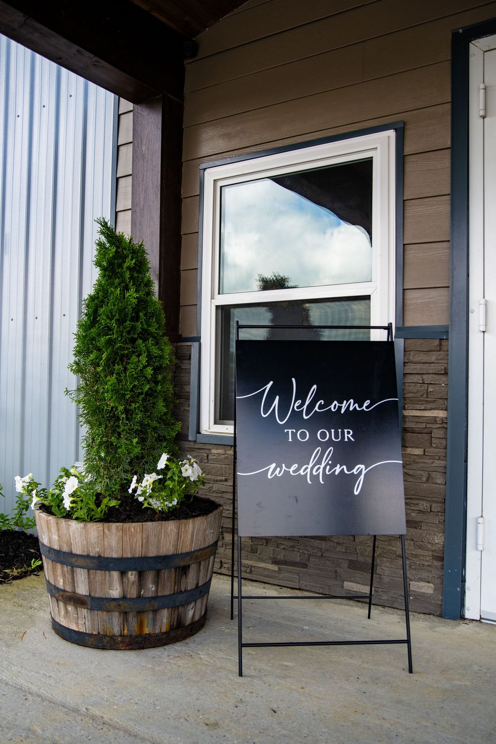 Elegant black wedding welcome sign on easel beside barrel planter with white flowers and evergreen shrub at venue entrance.