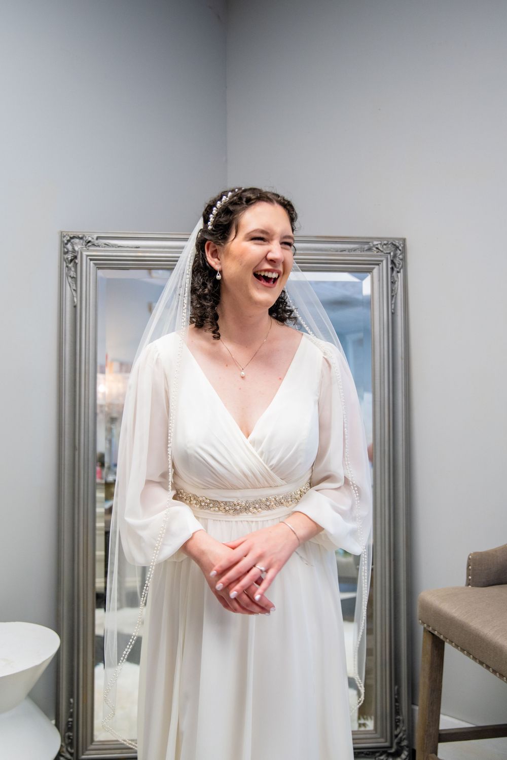 Radiant bride in white chiffon gown with jeweled belt and veil laughing joyfully in a bridal boutique fitting room.