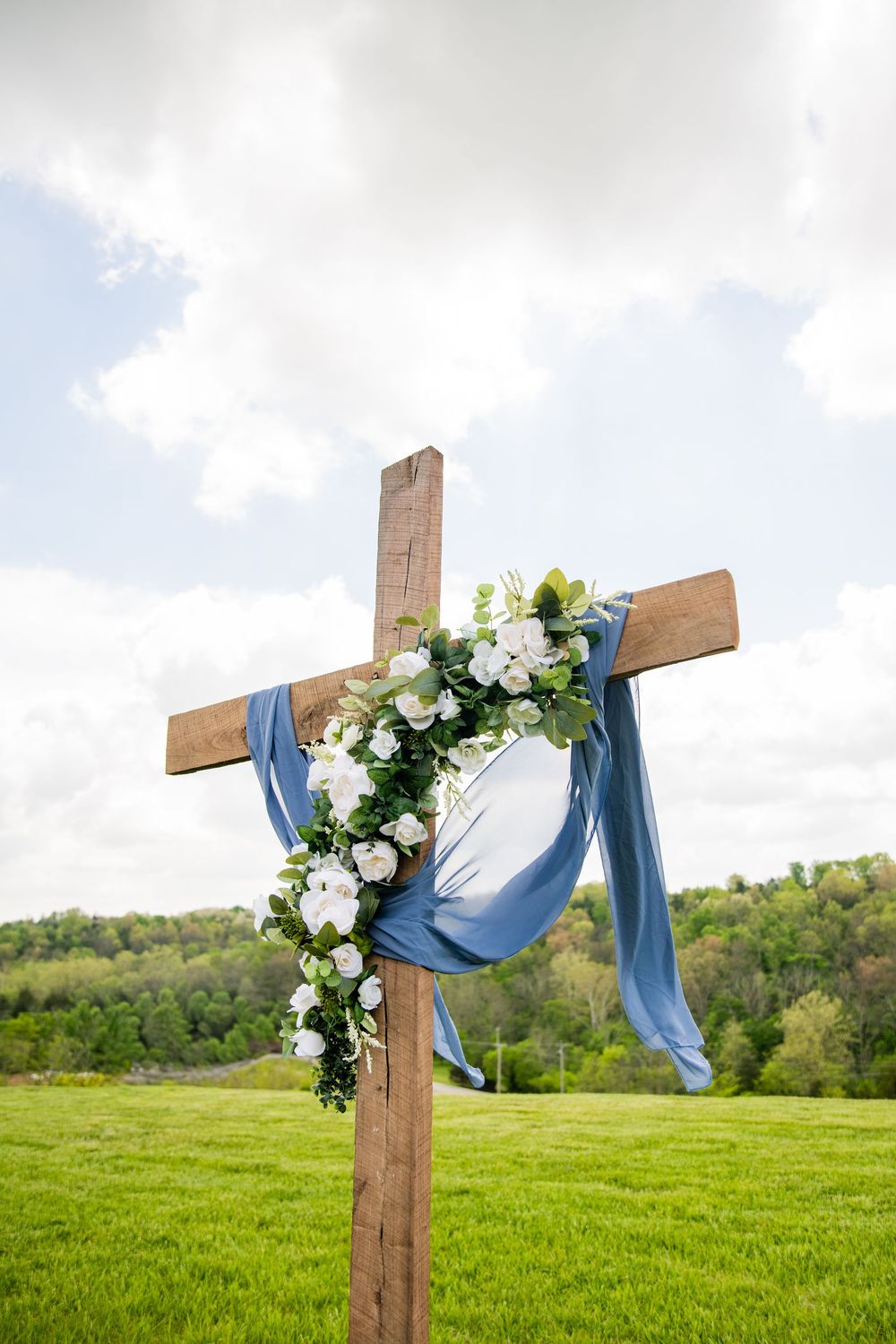 Rustic wooden cross decorated with white flowers and blue fabric draped over it stands in a green field.