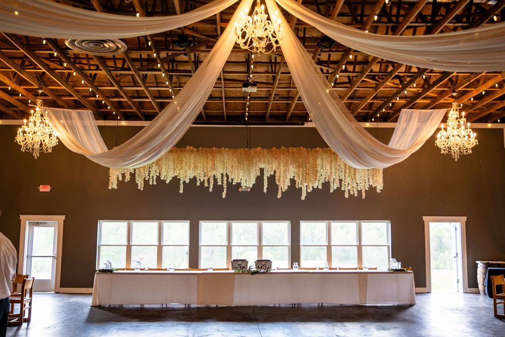 Elegant barn wedding venue with draped white fabric, crystal chandeliers, string lights, and a decorated head table.