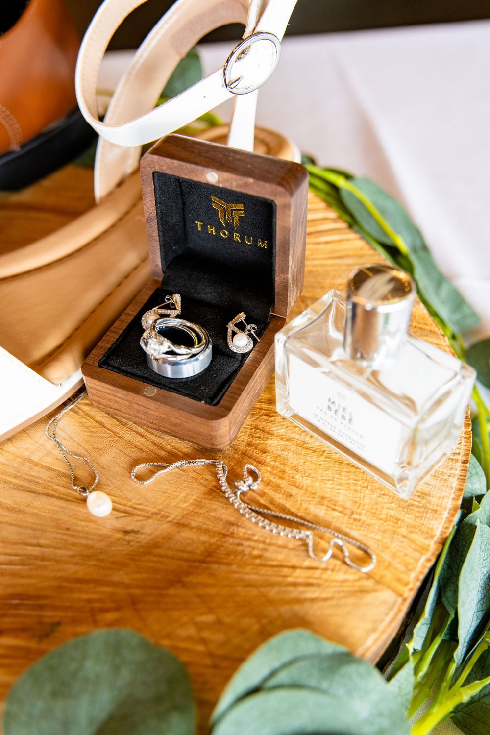 Wedding accessories including rings in Thorum box, perfume bottle, and eucalyptus on wood surface.