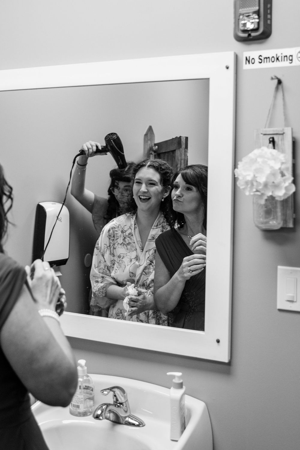 Bridesmaids getting ready together, styling hair in a bathroom mirror, black and white photo.