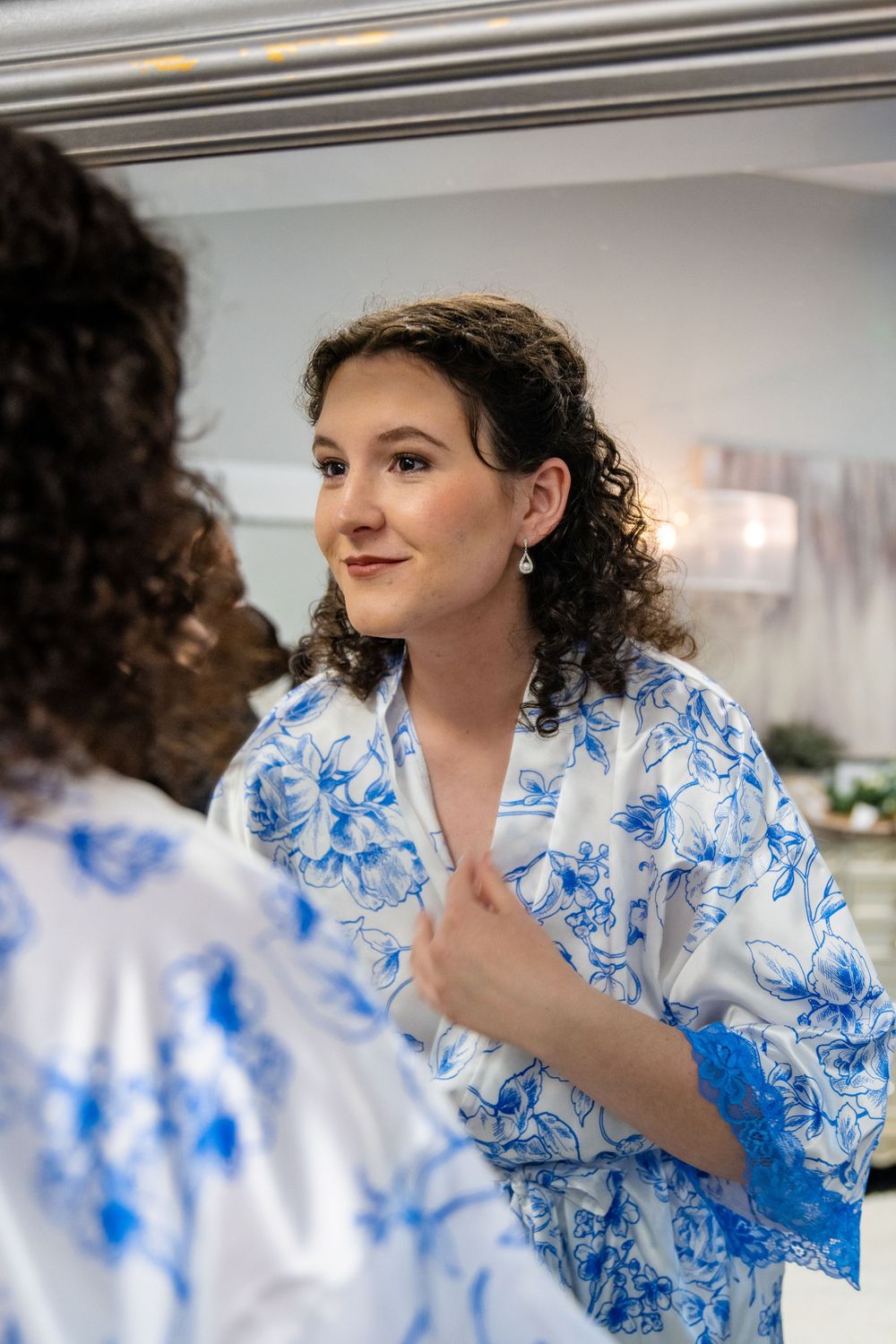 Bride in blue floral robe smiling at reflection in mirror while getting ready for wedding day.