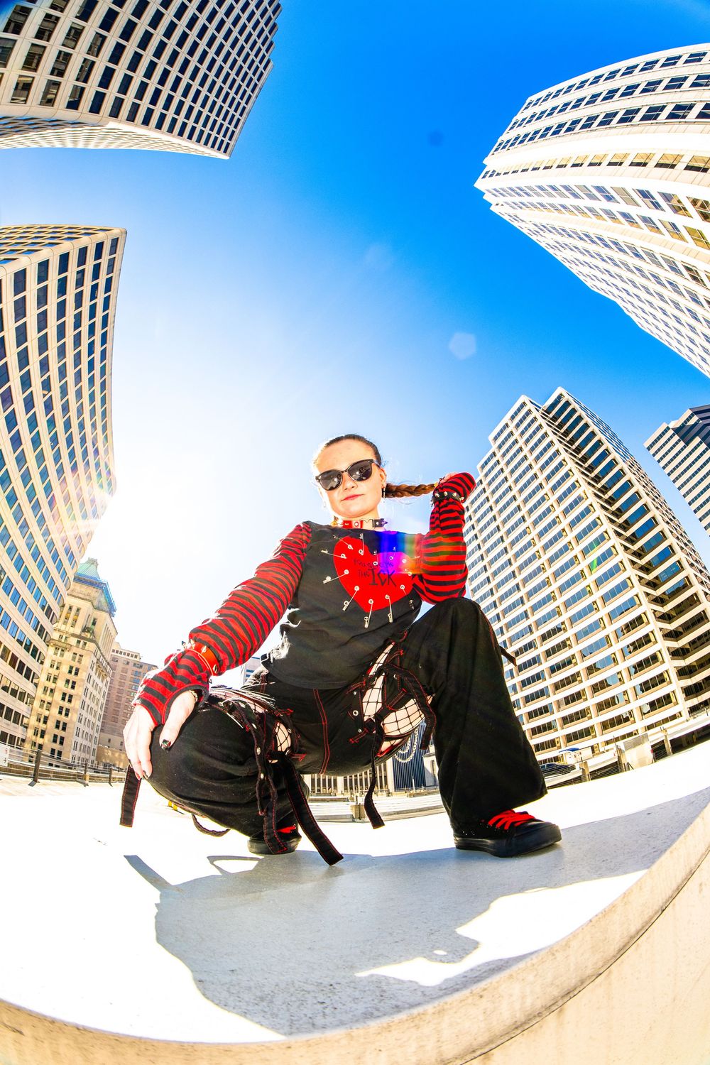 A punk-styled person sits confidently outdoors surrounded by towering skyscrapers under a bright blue sky, shot with a fisheye lens.