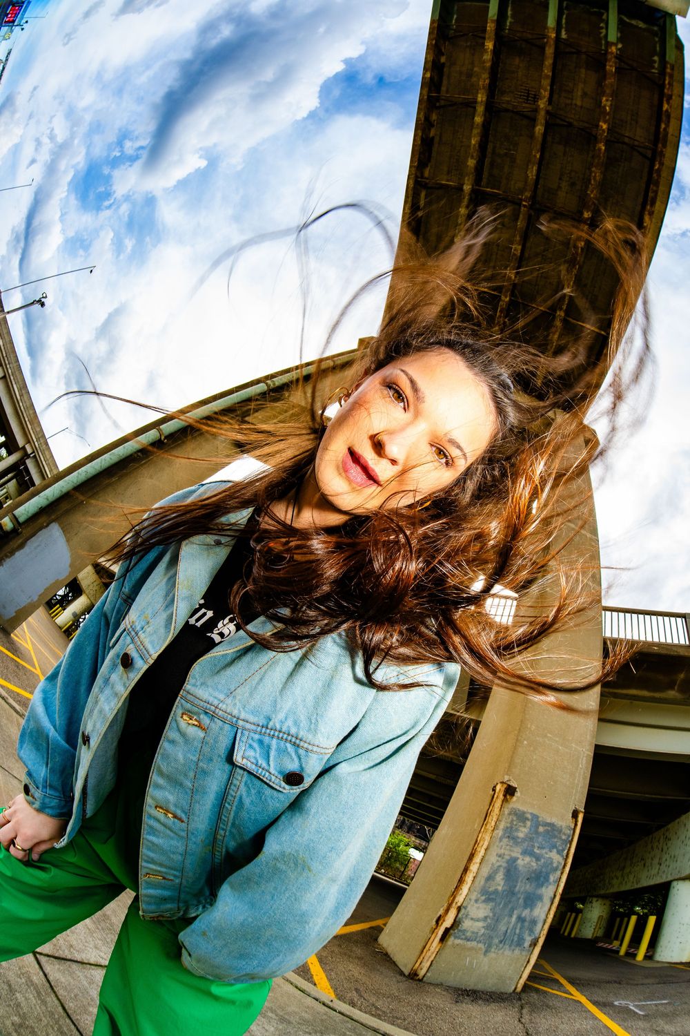 Young woman in denim jacket and green pants with wind-blown hair posing under a highway overpass.
