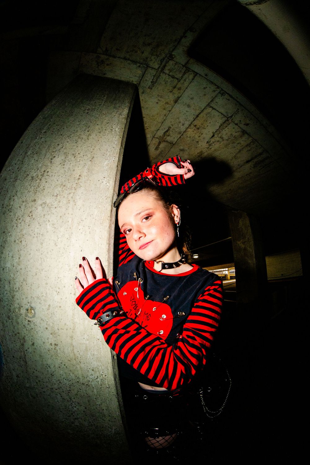 Young woman in red and black striped gothic outfit posing dramatically in dark concrete tunnel with fisheye lens effect.
