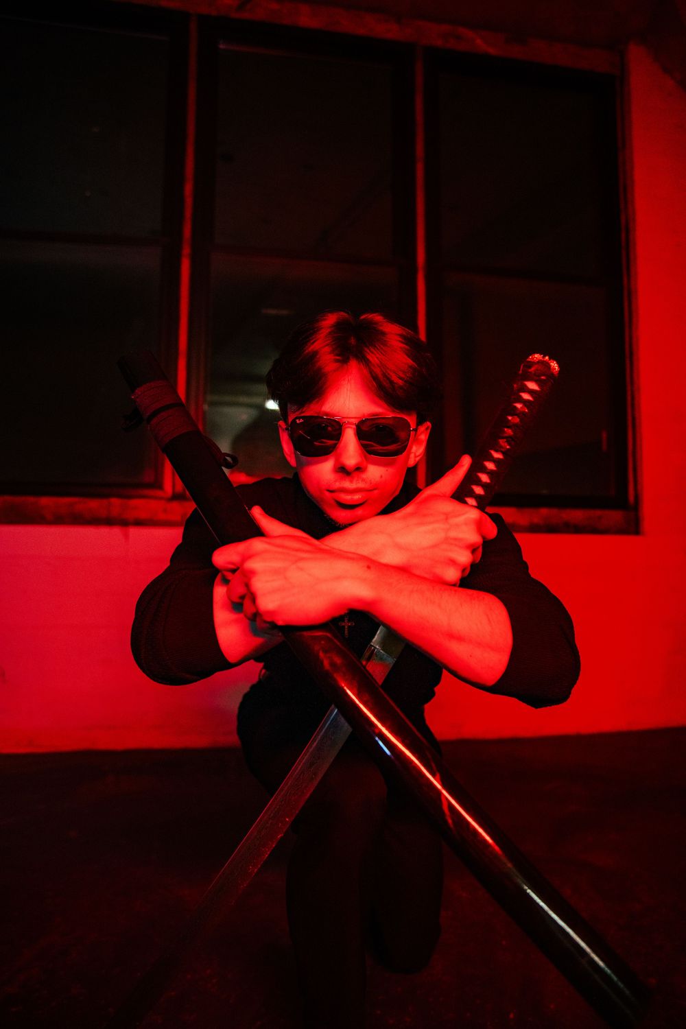 Man in black holding crossed swords under red neon lighting in dramatic pose.
