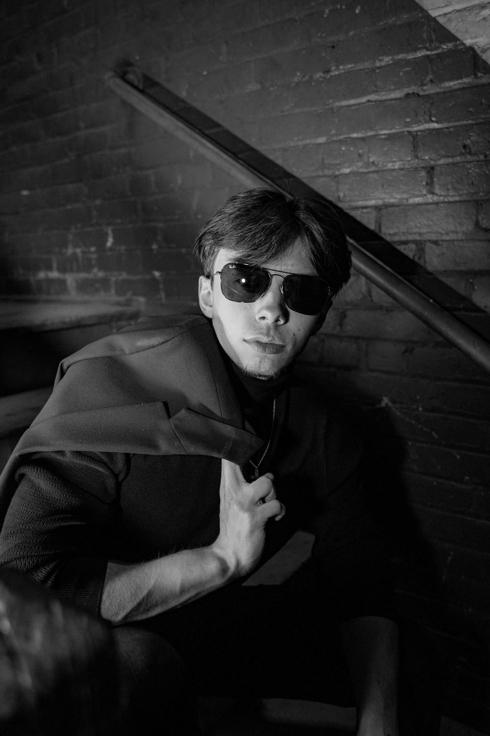 Man in black outfit and aviator sunglasses posing on stairs against brick wall in black and white photo.